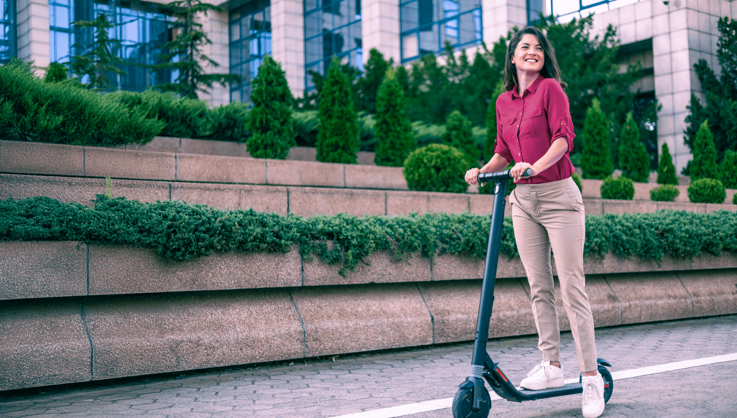 a young woman with a scooter passing in front of a building with greenery
