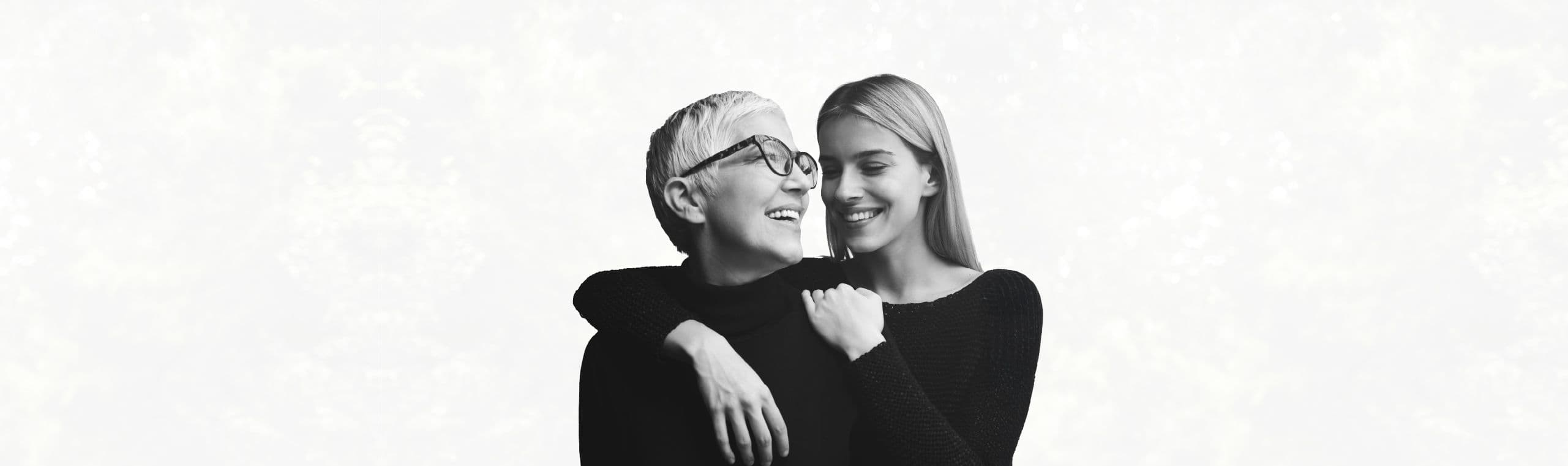 A visibly younger woman is hugging a white-haired woman from behind, both smiling and looking close