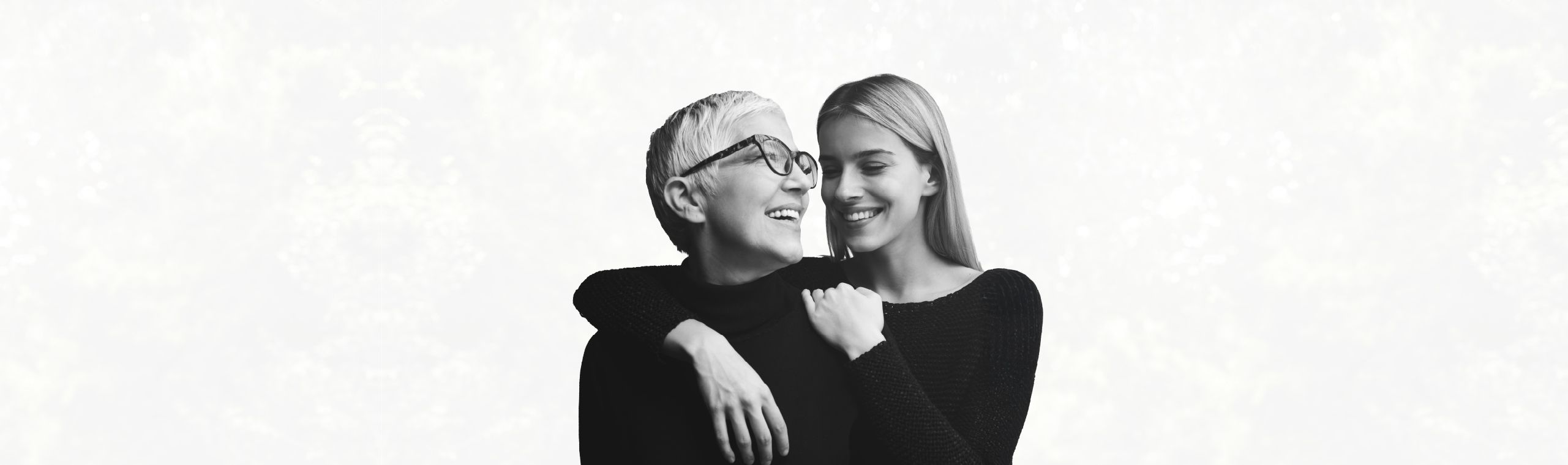 A visibly younger woman is hugging a white-haired woman from behind, both smiling and looking close
