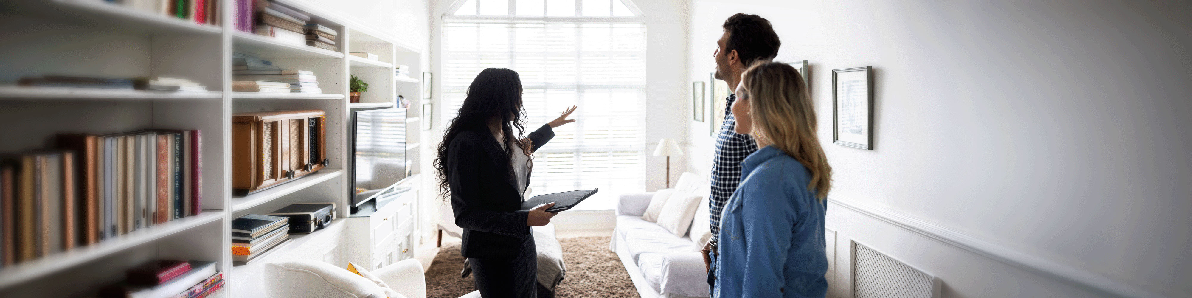 Real estate agent gesturing toward a large window while showing a modern living room to a couple