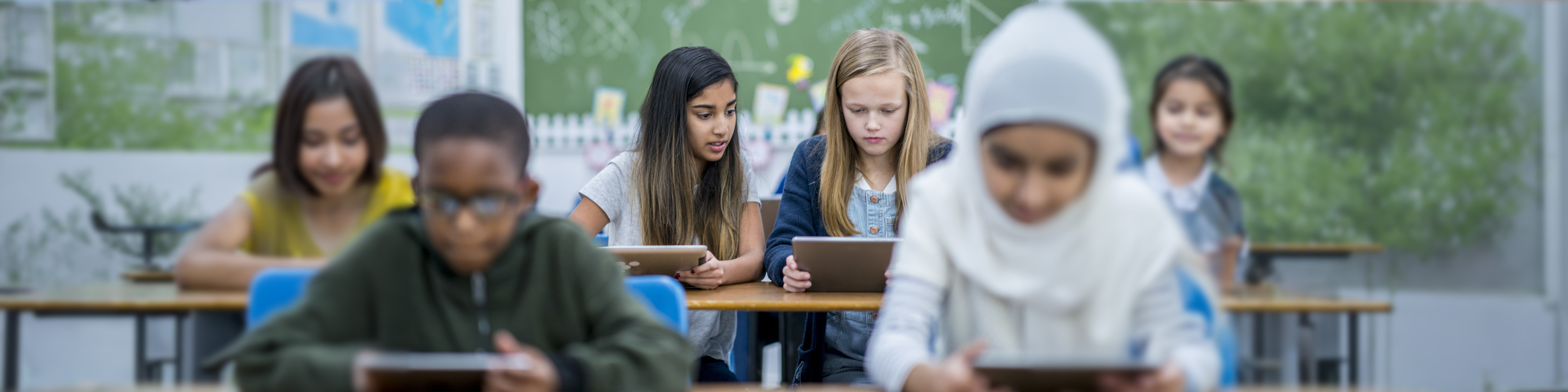 EN: Schoolchildren from diverse backgrounds using tablets in a classroom, including a girl wearing a headscarf in the foreground. FR: Élèves de différents horizons utilisant des tablettes en classe, avec une fille portant un foulard au premier plan.